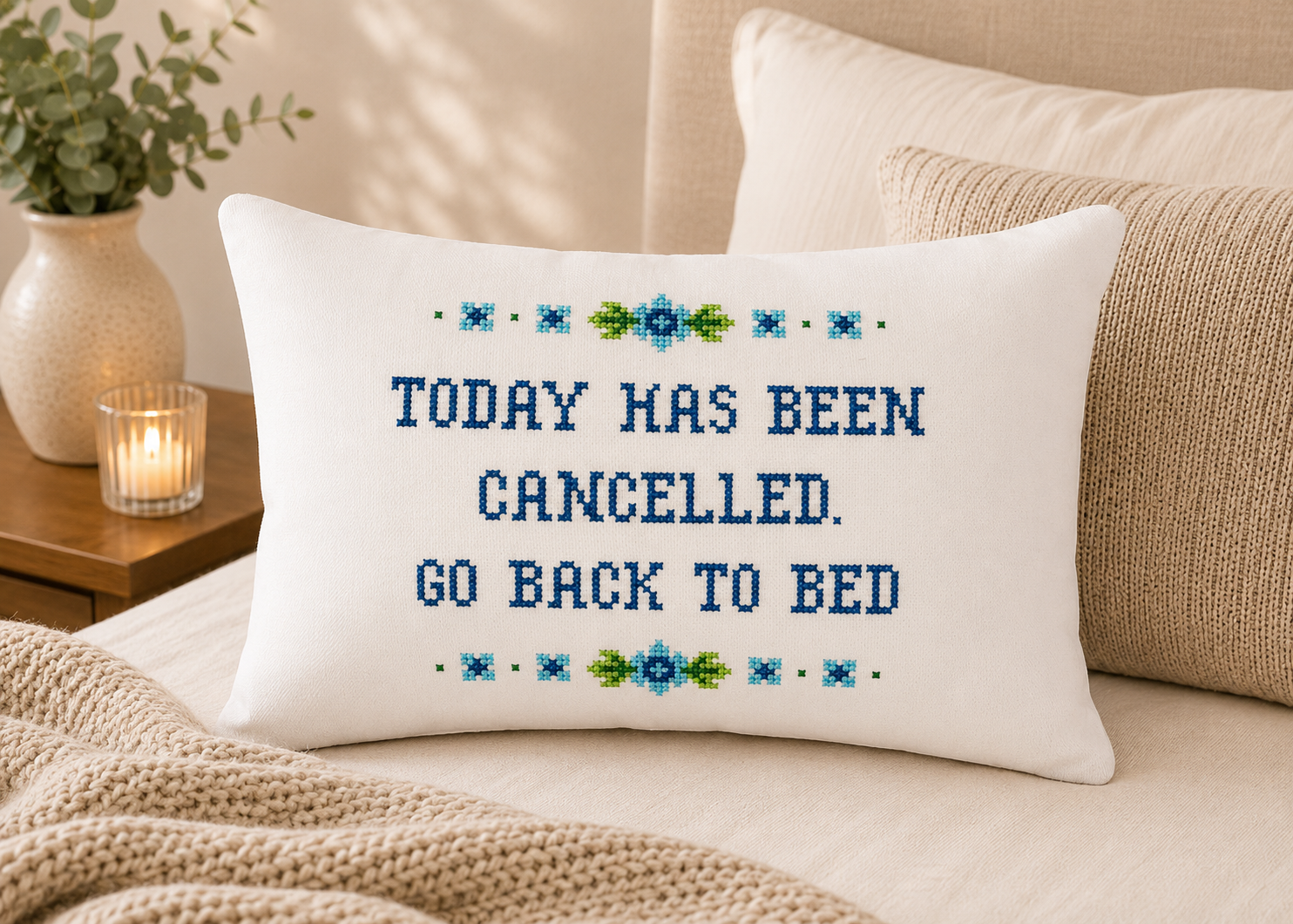 Decorative pillow with cross-stitched text on a couch, surrounded by pillows and a candle.