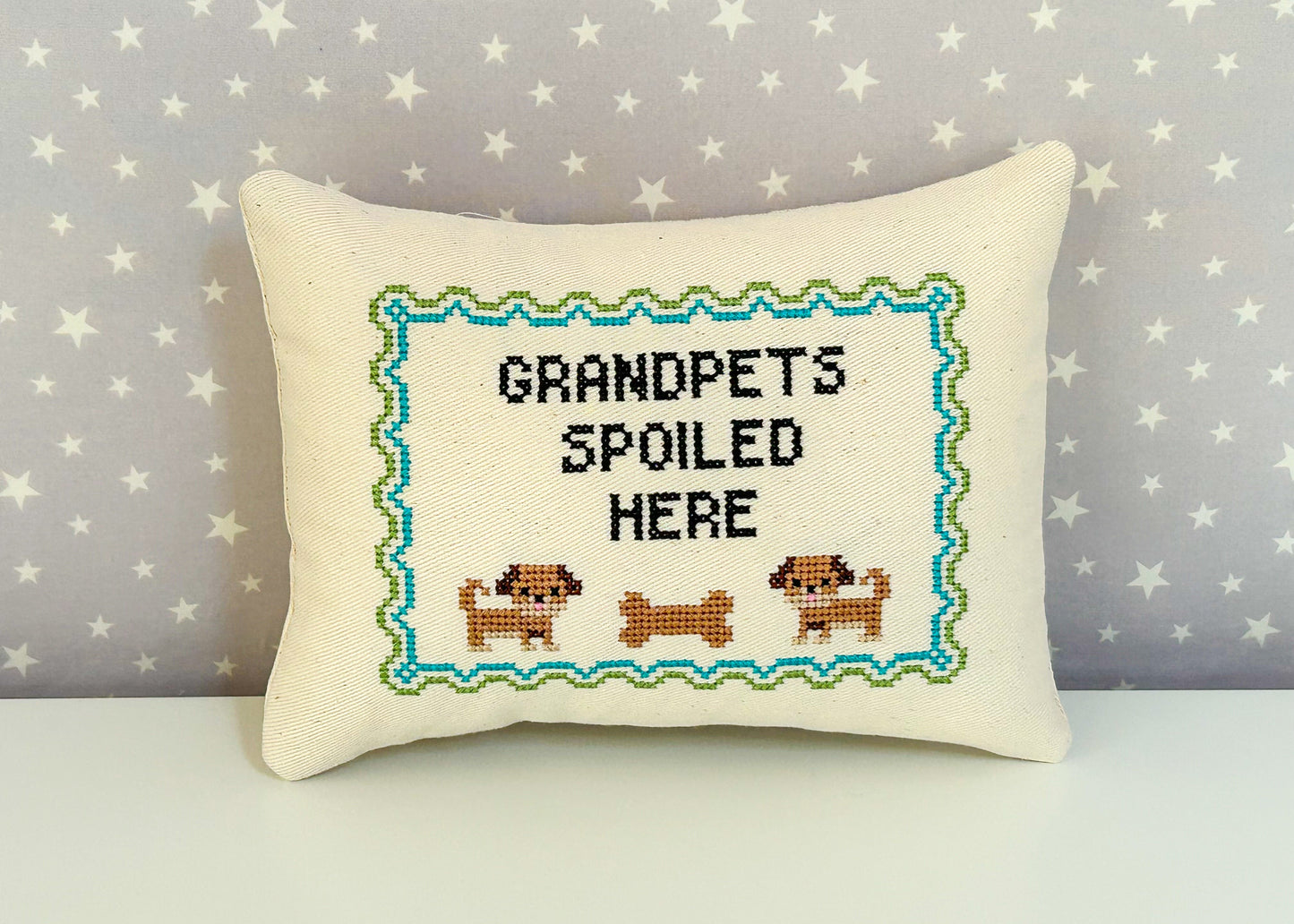 Decorative pillow with cross-stitched design and text 'Grandpets Spoiled Here' on a star-patterned background.
