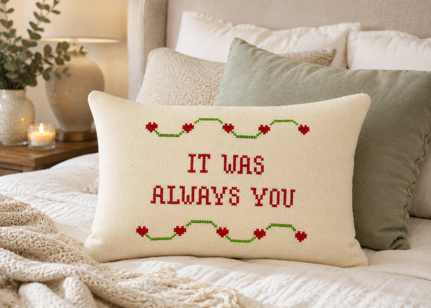 Decorative pillow with 'It was always you' text on a bed with pillows and a lamp in the background.