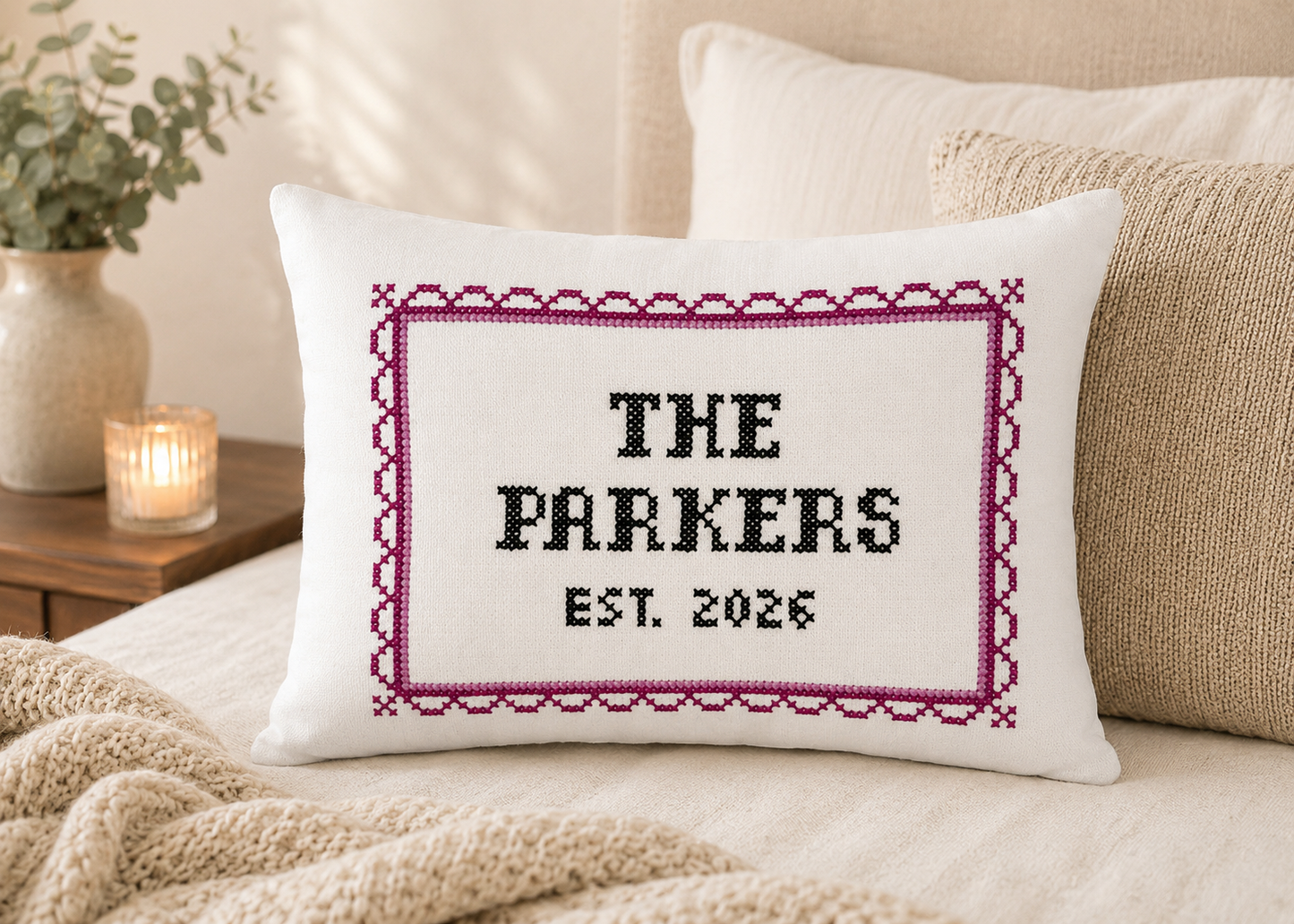 Decorative pillow with cross-stitched text on a couch, surrounded by pillows and a candle.