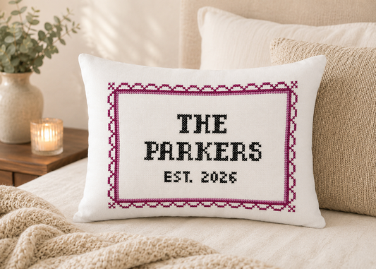 Decorative pillow with cross-stitched text on a couch, surrounded by pillows and a candle.