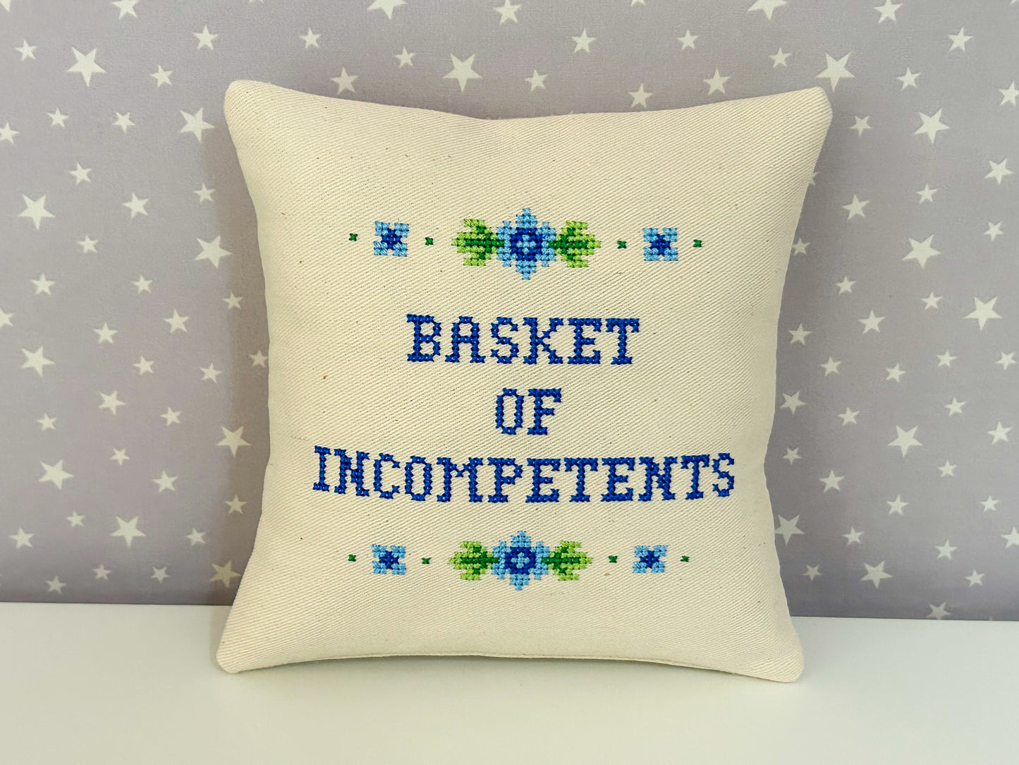 Decorative pillow with cross-stitched text 'BASKET OF INCOMPETENTS' on a star-patterned background