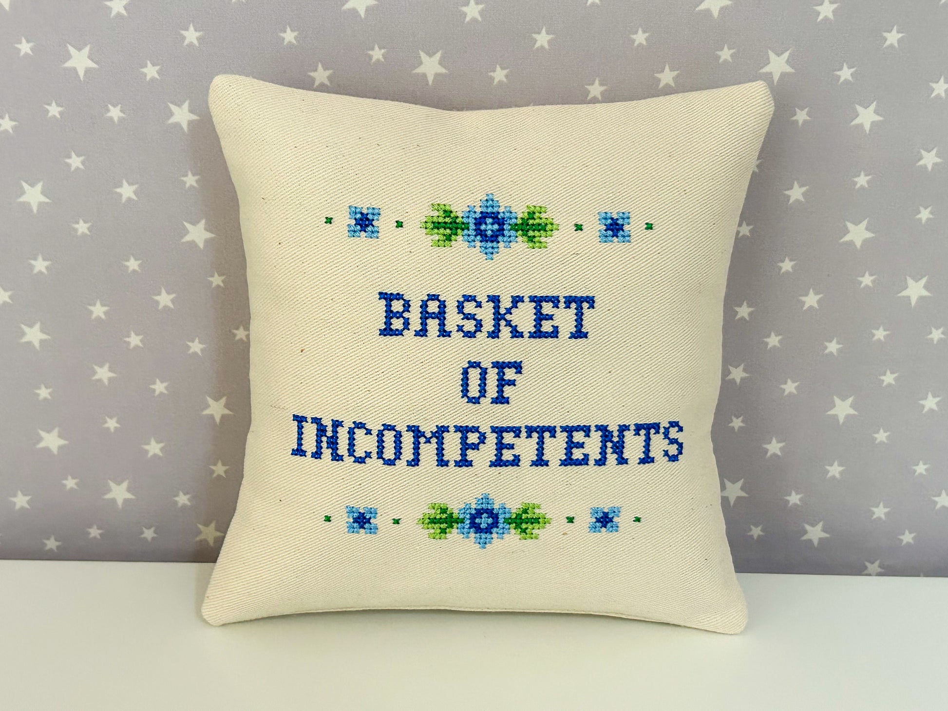Decorative pillow with cross-stitched text 'BASKET OF INCOMPETENTS' on a star-patterned background