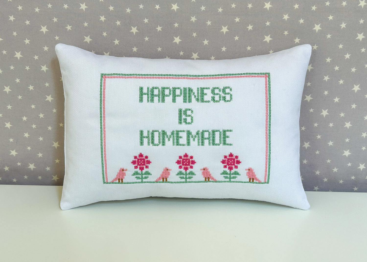 White pillow with the text happiness is homemade stitched using cross stitching with a border of birds and flowers. Grey background with stars