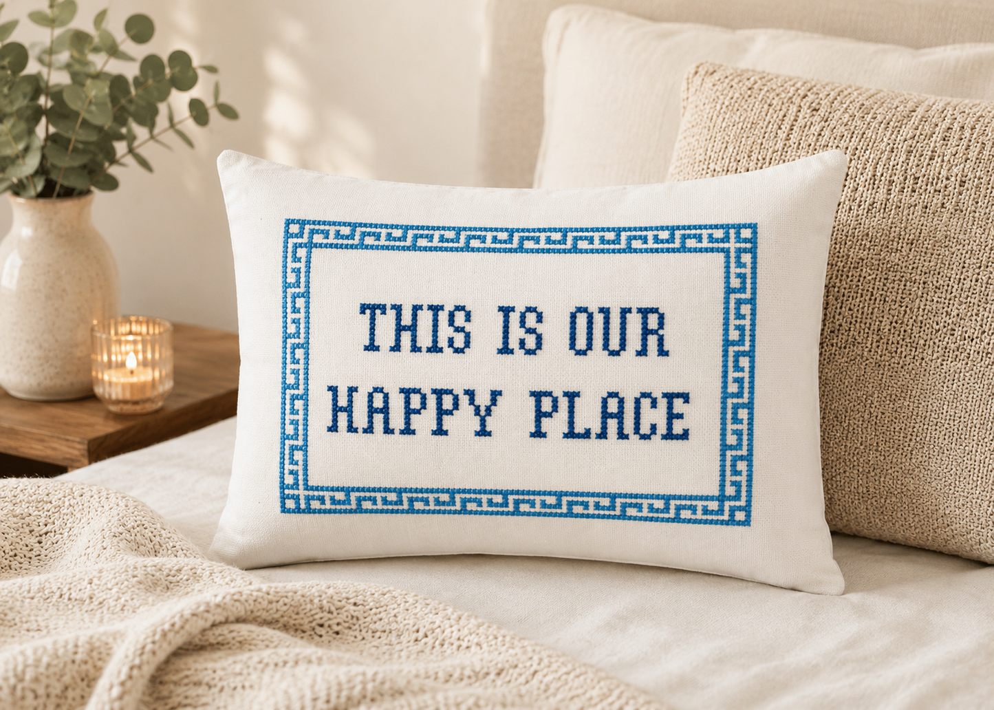 Decorative pillow with cross-stitched text on a bed, surrounded by pillows and a candle.