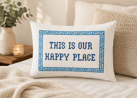 Decorative pillow with cross-stitched text on a bed, surrounded by pillows and a candle.